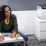 Imprimante Xerox® VersaLink® C620 dans un bureau Femme souriante dans un bureau à côté de l'imprimante couleur Xerox® VersaLink® C620
