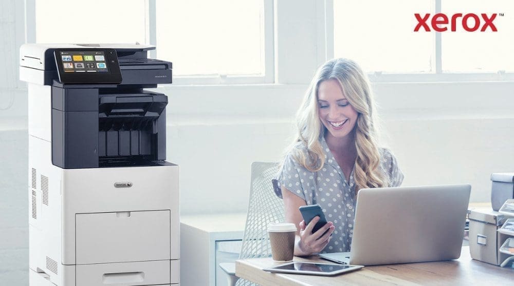 Une personne regarde joyeusement son smartphone, assise à un bureau avec un ordinateur portable. Une imprimante multifonction Xerox VersaLink B605 est visible à côté du bureau, dans un espace de travail lumineux.
